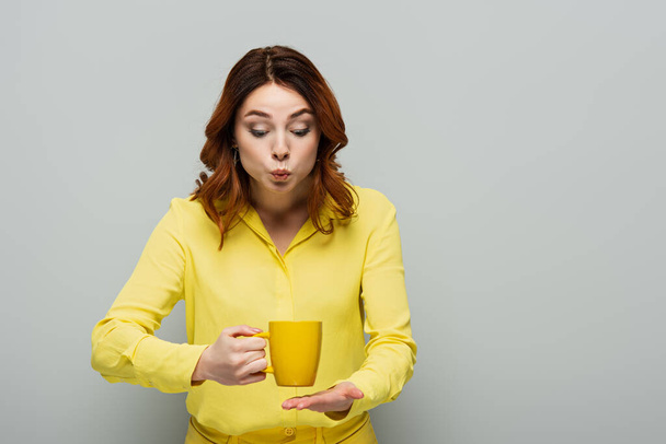 шокована жінка в жовтій блузці тримає чашку гарячого чаю на сірому
 - Фото, зображення