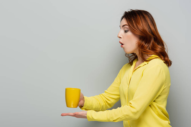 шокована жінка в жовтій блузці тримає чашку гарячого напою на сірому
 - Фото, зображення