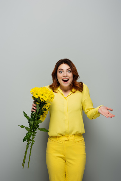 Erstaunte Frau mit gelben Blumen, während sie mit offenem Mund auf grau steht - Foto, Bild