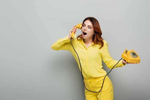 mujer excitada en ropa amarilla hablando por teléfono vintage mientras mira hacia otro lado en gris - Foto, Imagen