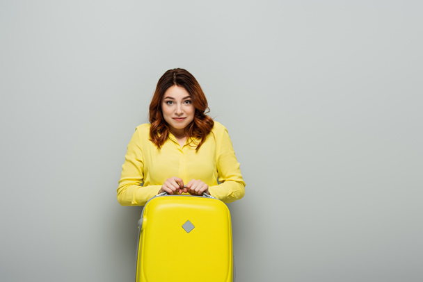 cheerful woman in yellow blouse looking at camera while standing with suitcase on grey - Φωτογραφία, εικόνα