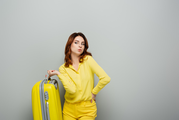 mujer reflexiva sosteniendo maleta amarilla mientras está de pie con la mano en la cadera en gris - Foto, Imagen