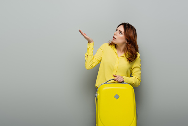 shocked woman pointing with hand while holding yellow suitcase on grey - Foto, imagen