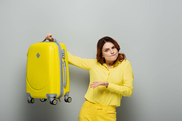 обескураженная женщина, указывающая на желтый чемодан, глядя в камеру на сером - Фото, изображение