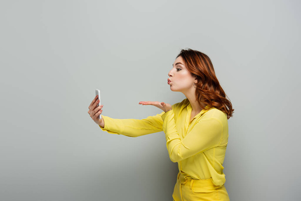 žena s vlnitými vlasy foukání polibek při užívání selfie na smartphone na šedé - Fotografie, Obrázek
