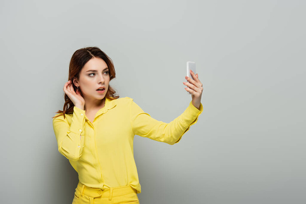 flirty woman adjusting hair while taking selfie on cellphone on grey - Photo, Image