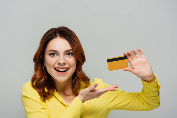 fröhliche Frau blickt in die Kamera und zeigt auf Kreditkarte isoliert auf grau - Foto, Bild
