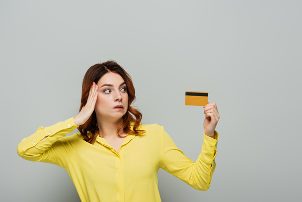 bemused woman touching head while holding credit card on grey - Foto, imagen