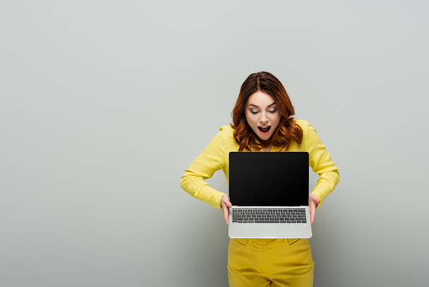 amazed woman with curly hair holding laptop with blank screen on grey - Photo, Image