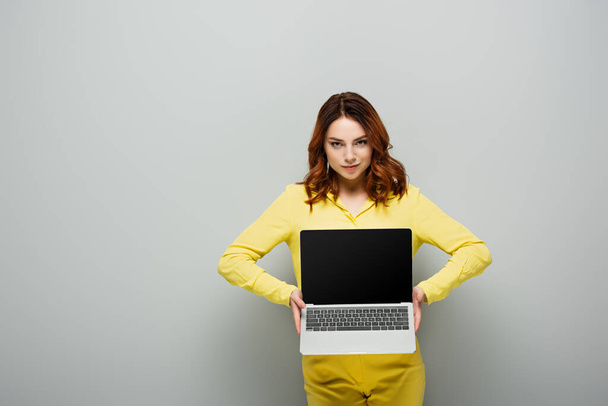 grijnzende vrouw op zoek naar camera terwijl het houden van laptop met leeg scherm op grijs - Foto, afbeelding