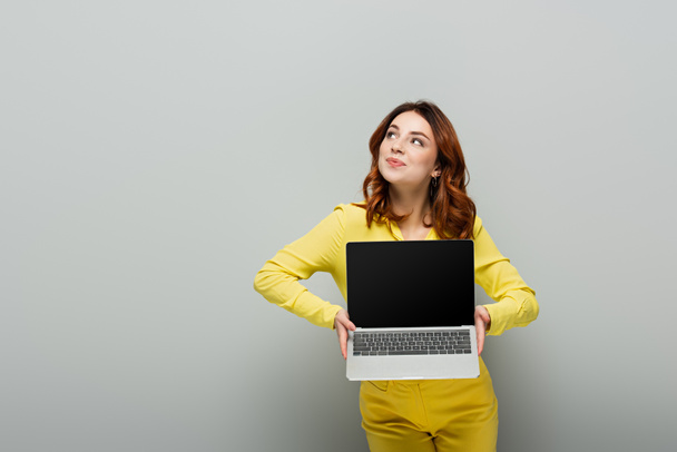 smiling woman looking away while holding laptop with blank screen on grey - Foto, imagen