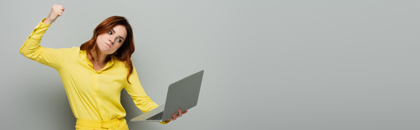 ärtynyt nainen, jolla kannettava tietokone seisoessaan puristi nyrkkiä harmaalla, banneri - Valokuva, kuva