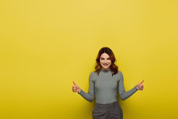 szczęśliwa kobieta z kręconymi włosami pokazując kciuki w górę na żółty - Zdjęcie, obraz