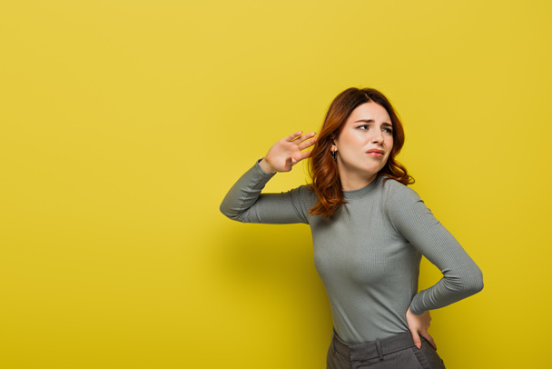 displeased woman with curly hair standing with hand on hip while listening on yellow - Photo, Image