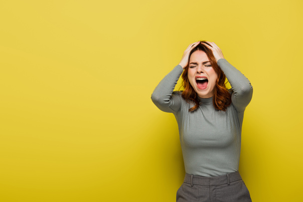 Wütende Frau mit welligem Haar schreit und berührt Kopf auf gelb  - Foto, Bild