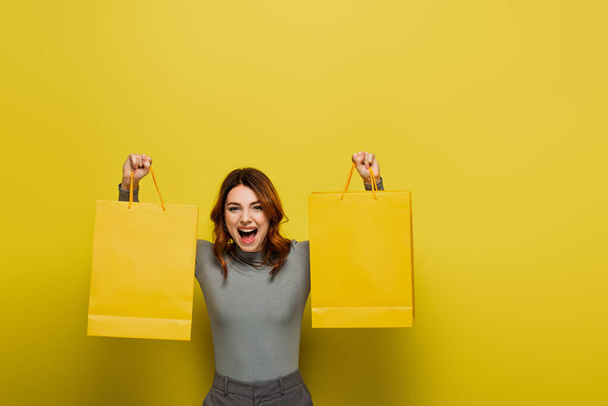 verbaasd jonge vrouw met krullend haar houden shopping tassen op geel - Foto, afbeelding