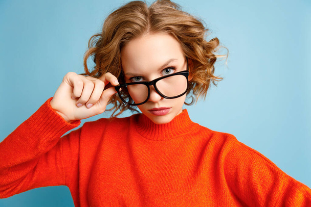 Femme curieuse en pull rouge ajustant les lunettes et regardant de près la caméra sur fond bleu - Photo, image