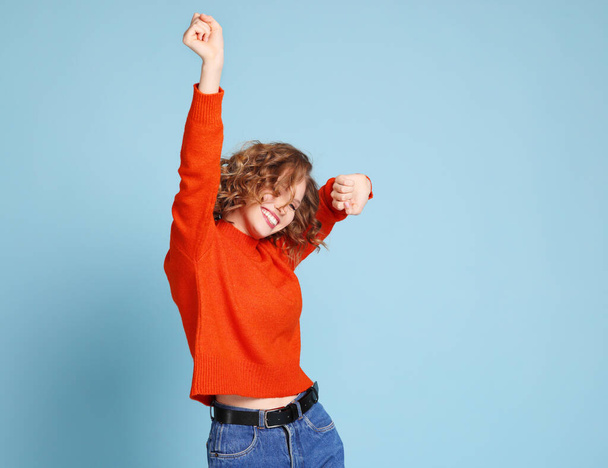 Happy woman in orange sweater smiling and dancing energetically with closed eyes against blue background - 写真・画像