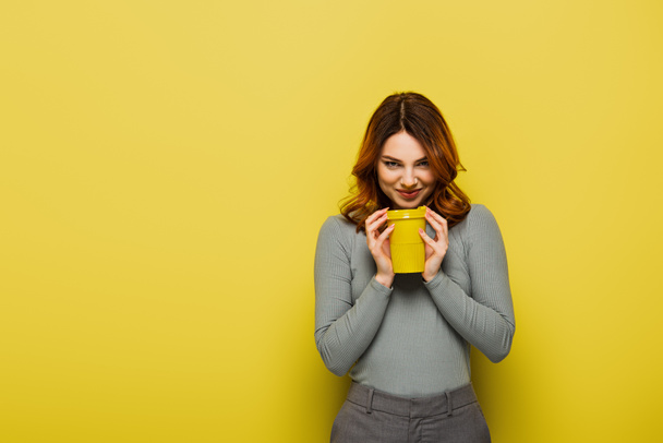 listige junge Frau mit lockigem Haar hält Pappbecher in der Hand und lächelt auf gelb - Foto, Bild