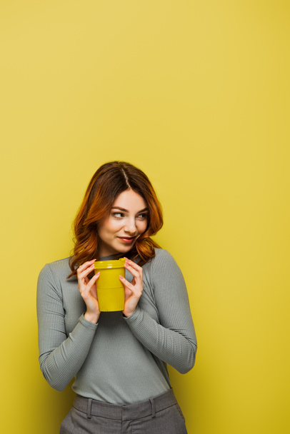joyful young woman with curly hair holding paper cup and looking away on yellow - Фото, изображение