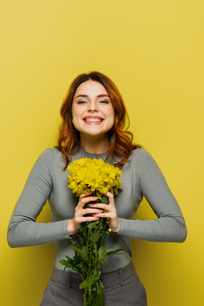 Χαρούμενη νεαρή γυναίκα με κυματιστά μαλλιά που κρατάει λουλούδια στο κίτρινο - Φωτογραφία, εικόνα