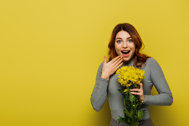 donna stupita con i capelli ondulati che tengono fiori sul giallo - Foto, immagini