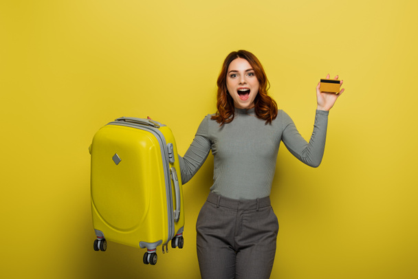 verblüffte Frau mit lockigem Haar, die Gepäck und Kreditkarte auf gelb hält - Foto, Bild