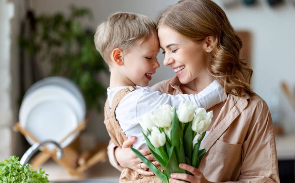 Optimistic family: mother with bouquet of  tulips smiling and touching forehead with happy son  during holiday celebration  mothers day at home - Foto, imagen