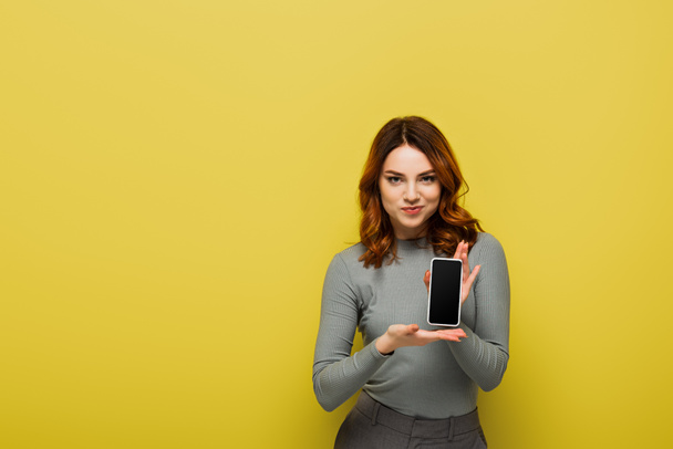 mujer alegre con pelo rizado sosteniendo teléfono inteligente con pantalla en blanco en amarillo - Foto, Imagen