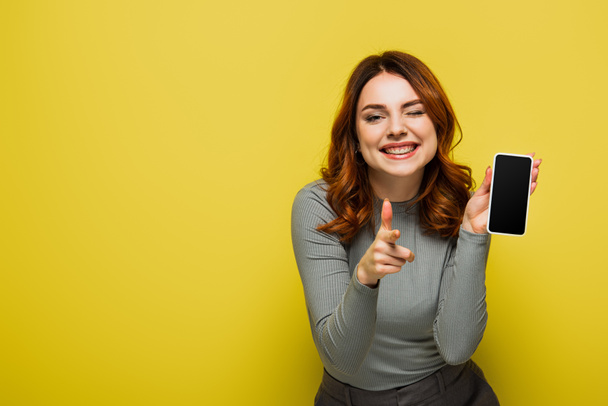 femme gaie tenant smartphone avec écran vide et pointant du doigt tout en regardant la caméra sur jaune - Photo, image