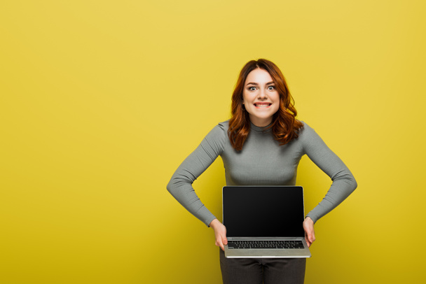 fröhliche junge Frau mit lockigem Haar hält Laptop mit leerem Bildschirm und beißenden Lippen auf gelb - Foto, Bild