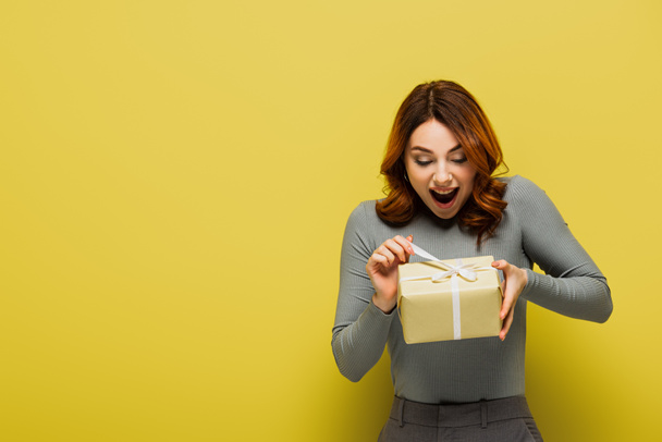 amazed young woman pulling ribbon on present on yellow - Photo, Image
