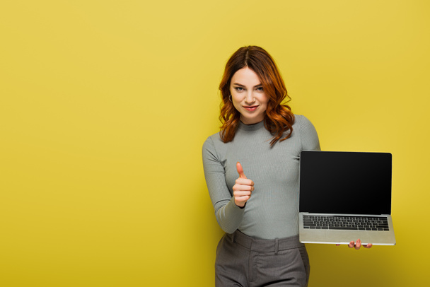 ευτυχισμένη γυναίκα με σγουρά μαλλιά κρατώντας φορητό υπολογιστή με κενή οθόνη, ενώ δείχνει τον αντίχειρα επάνω στο κίτρινο - Φωτογραφία, εικόνα