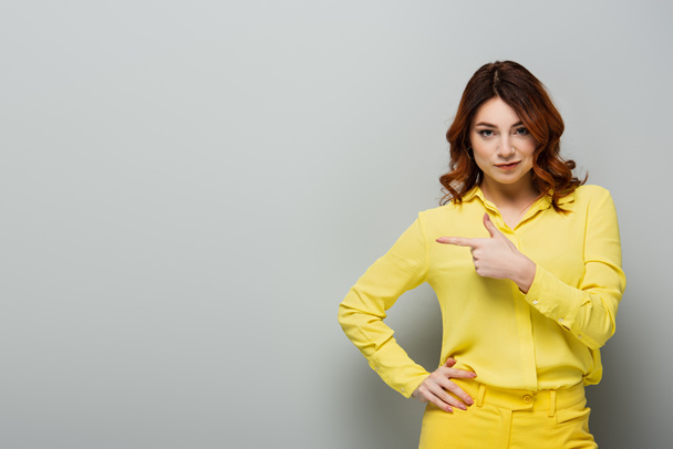 νεαρή σγουρή γυναίκα σε κίτρινο πουκάμισο στέκεται με το χέρι στο ισχίο και δείχνοντας με το δάχτυλο σε γκρι  - Φωτογραφία, εικόνα