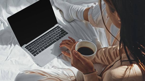 korkea kulma näkymä nainen tilalla kuppi kahvia lähellä kannettavaa tietokonetta tyhjä näyttö sängyllä - Valokuva, kuva
