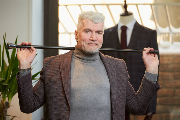 A smiling mature man with gray hair and a sporty physique is holding a fiber carbon cane with two hands behind his head in a clothing store. A male customer with a beard wears a suit in a boutique. - Photo, Image