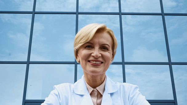 Glimlachende arts in witte jas kijkend naar camera in de buurt van gebouw  - Foto, afbeelding