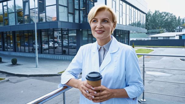 Smiling doctor in white coat holding coffee to go outdoors  - Foto, Bild
