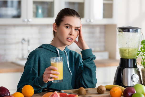 young woman touching head while holding fresh smoothie near fruits in kitchen - Photo, Image