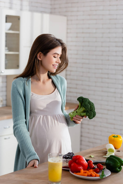 alegre mujer embarazada sosteniendo brócoli mientras se prepara el desayuno en la cocina  - Foto, Imagen