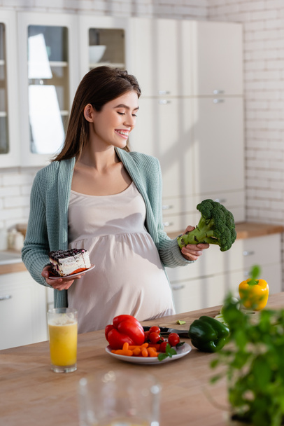 χαρούμενη έγκυος γυναίκα επιλέγοντας ανάμεσα σε κέικ και φρέσκο λάχανο στην κουζίνα - Φωτογραφία, εικόνα