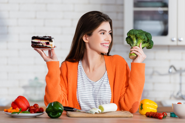 mujer alegre sosteniendo pastel y brócoli fresco cerca de verduras en la cocina - Foto, Imagen