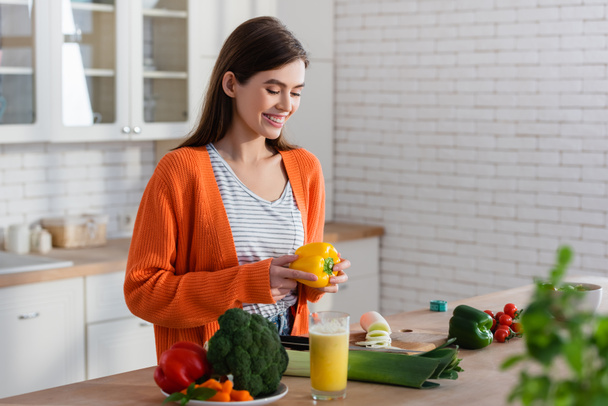 Улыбающаяся женщина держит перец рядом с овощами на кухонном столе, размытый передний план - Фото, изображение