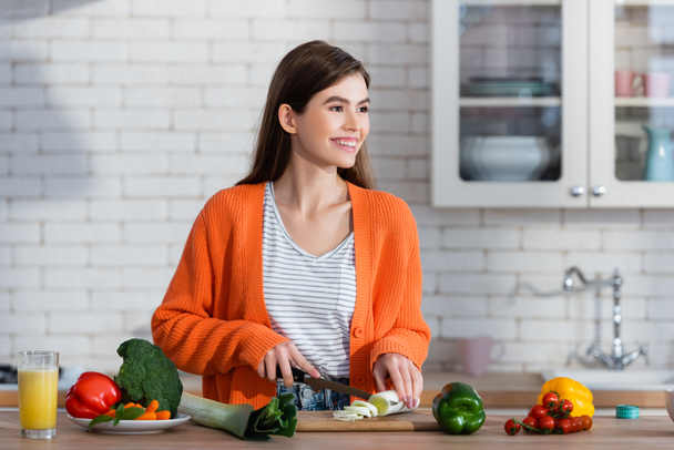 mujer alegre mirando hacia otro lado mientras corta puerro fresco cerca de verduras en la mesa - Foto, Imagen