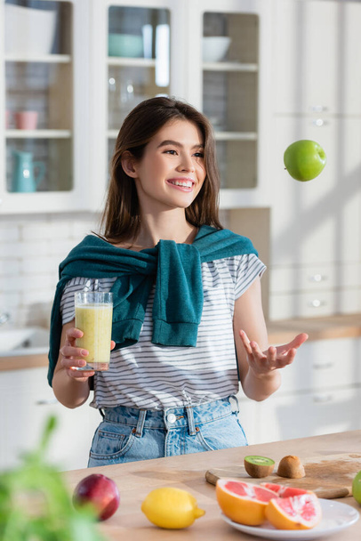 cheerful woman juggle with apple while holding fresh smoothie on blurred foreground - Photo, Image