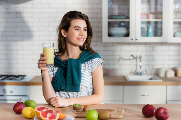 sonriente mujer sosteniendo jugo fresco cerca de frutas en la mesa de la cocina - Foto, Imagen