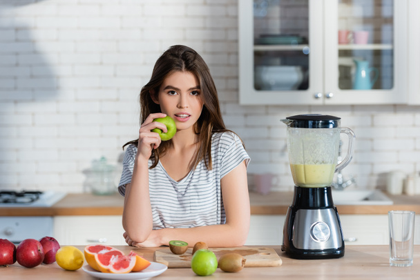 молодая женщина смотрит в камеру, держа яблоко возле свежих фруктов и блендера - Фото, изображение