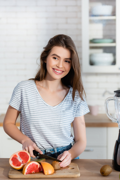 cheerful woman looking away while cutting fresh fruits in kitchen - Photo, Image
