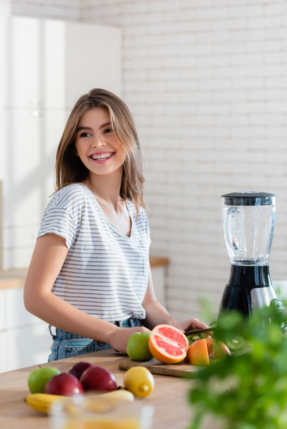 cheerful woman looking away while preparing fruit breakfast on blurred foreground - Photo, Image