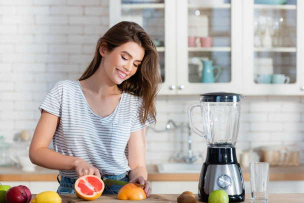 szczęśliwa kobieta cięcia świeżych owoców w pobliżu blendera w kuchni - Zdjęcie, obraz